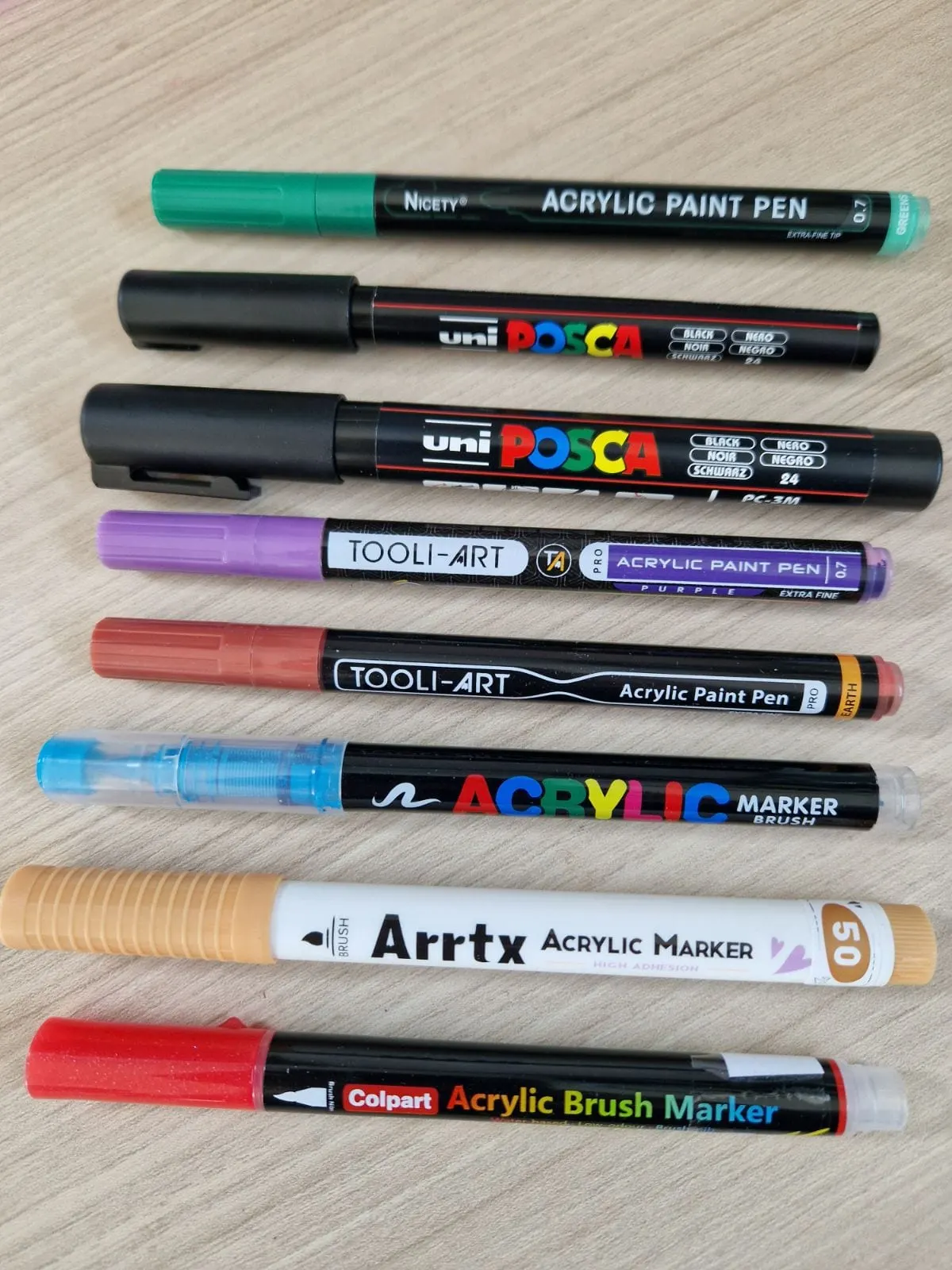 Plusieurs marqueurs acryliques de différentes marques (Posca, Nicety, Tooli-art, Arrtx, Colpart, GuangNa) alignés.